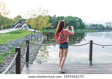 woman in summer in a city park near a reservoir, a pond, river and lake, records video photos on a smartphone. Sightseeing and travel. Back view. Denim shorts jacket with backpack