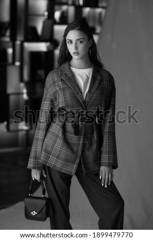 Elegant girl in red blazer, brown wool trousers standing and posing against textile background and interior with lights. Pretty young woman with natural makeup and long black straight hair.