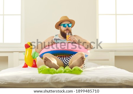 Funny young man in sunglasses and summer wear sipping beach cocktail, sitting barefoot in inflatable lifebuoy at home. Covid-19 quarantine, vacation in lockdown, canceled holiday travel plans concept Royalty-Free Stock Photo #1899485710