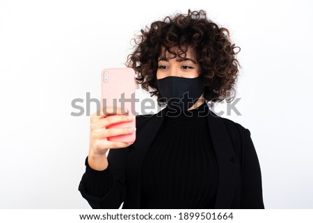 Isolated shot of pleased cheerful young beautiful caucasian woman wearing medical mask standing against white wall, makes selfie with mobile phone. People, technology and leisure concept.