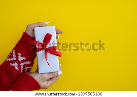 Female hands hold a white gift box with a red ribbon on a yellow paper background. Gift concept for Valentine's Day holiday or wedding