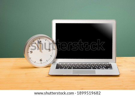Laptop with blank screen, clock with arrows at five to twelve on a wooden table. mockup for your text.distance learning concept