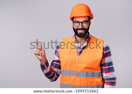 Cheerful Arab builder in hardhat and uniform pointing at copy space isolated over grey background