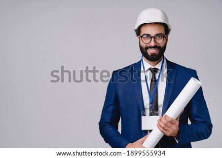 Cheerful young Arabian engineer with architectural plan and hardhat isolated over grey background