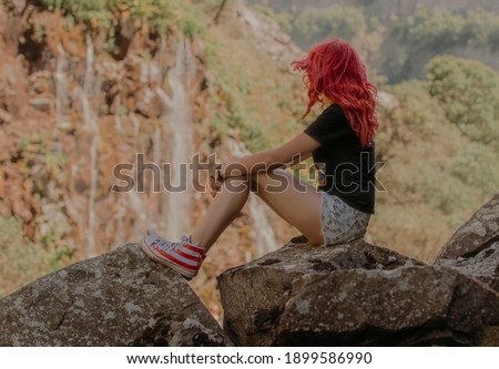 A beautiful girl with red hair in  shorts sits on a rock and watches the waterfall. The waterfall is blurred.