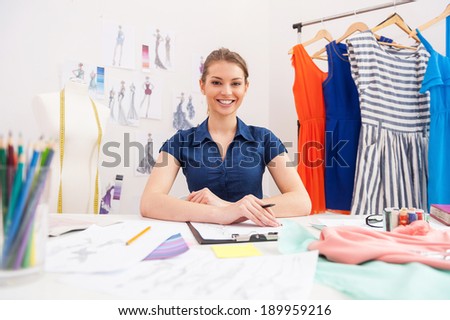 Confident fashion designer. Attractive female fashion designer looking at camera and smiling while sitting at her working place
