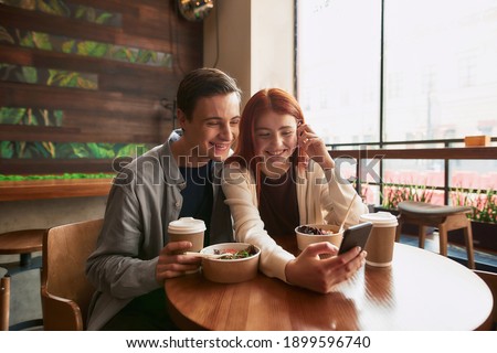 Happy teenage couple smiling while taking selfies using smartphone, sitting in a cafe together on a daytime. Modern relationships concept. Selective focus. Horizontal shot