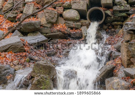 A torrent of water gushing from a concrete pipe high up in the side of a rocky hill on a rainy day. Dead leaves and branches on the ground.