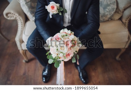 Wedding details groom. Stylish groom in a blue suit with a boutonniere sitting on the couch and holding a bouquet of close-ups.