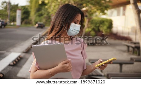   A beautiful woman with a mask to prevent the virus using her cell phone in the new normal, checking her social medias and carrying a laptop      