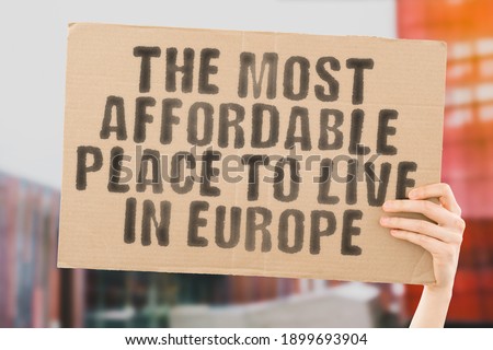 The phrase " The most affordable place to live in Europe " on a banner in men's hand with blurred background. Money. Earnings. Living. Salary. Apartment. Home. Expenses. Buying. Wealth