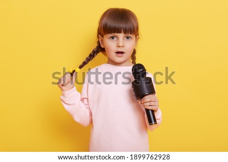 Shy small female kid with pigtails singing in microphone, cute little European girl wearing pale pink casual shirt looks at camera and performing, sings modern popular song, charming artist.
