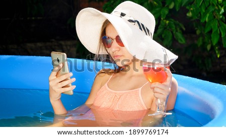 Beautiful smilin girl in summer hat and sunglasses relaxing in swimming pool and drinking cocktail while browsing social media on smartphone. Concept of happy summer holidays and vacation.