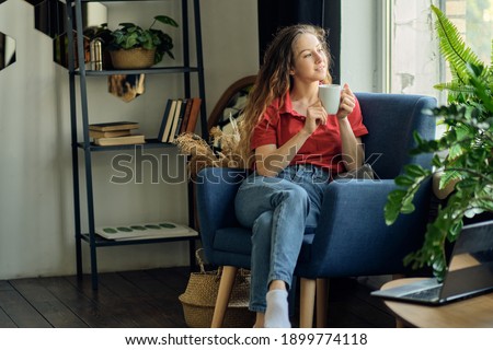 young woman with a cup of coffee in her hands sits in a chair by the window in a cozy living room