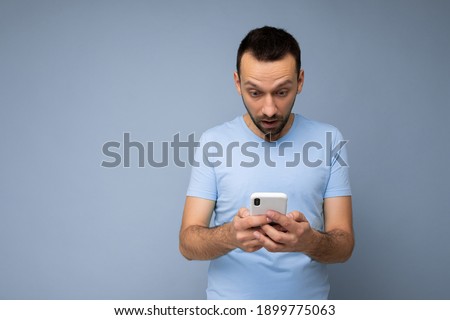 Photo of amazed handsome young man with beard wearing everyday blue t-shirt isolated over blue background holding and using mobile phone communication online on the internet looking at gadjet display