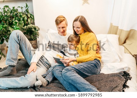 Happy family mother, father and boy at home in bed watch video in tablet. Green houseplants around, sustainable design. Yellow and gray colors 2021 year.