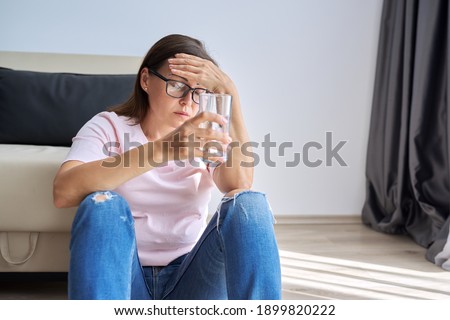 Sad middle aged woman sitting at home on the floor with glass of water. Health problems of older women, mental health, migraine pain, menopause symptom, depression period after coronavirus covid-19 Royalty-Free Stock Photo #1899820222