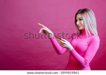 Woman with smartphone standing on the pink background and pointing her finger to the side. Woman over pink background with place for your text. High quality photo