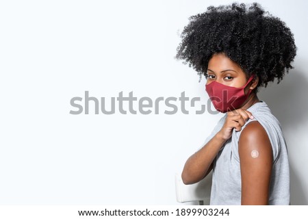 black teen girl shows the vaccine mark on her arm, wearing protective mask against covid-19, with a smile on her face, isolated on gray background Royalty-Free Stock Photo #1899903244