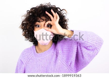 young beautiful caucasian woman wearing medical mask standing against white wall, doing ok gesture shocked with surprised face, eye looking through fingers. Unbelieving expression.