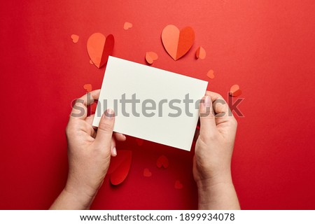 Blank greeting card or invitation. Female hands holding valentines day card mockup with paper hearts on red background