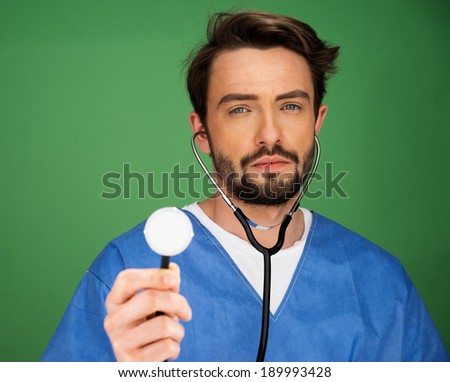 Handsome young male anaesthetist or doctor holding a stethoscope up with the disk facing the camera and the ear pieces in his ears as though he is listening, on a blue background