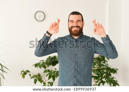 Photo of young man with beard in office crossing fingers and wishing