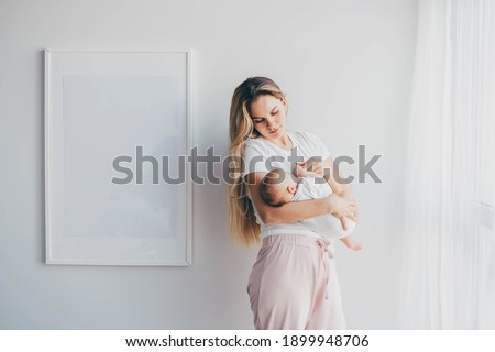 Careful mother with long loose flowing hair cradles baby holding daughter in arms and standing near large picture on wall under bright light at home.
