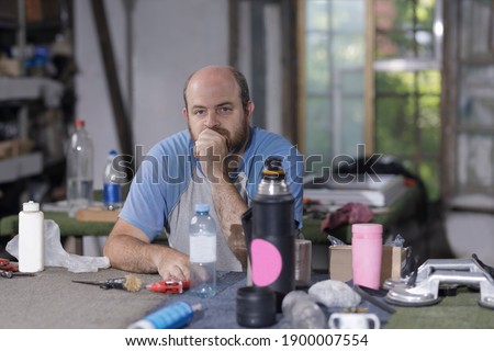 Caucasian man in a carpenter's workshop taking a break. Sitting at the work table, full of different elements, he looks into the camera. Disorderly atmosphere.