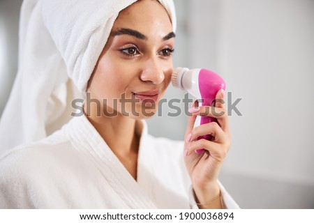 Close-up photo of hazel-eyed lady with towel wrapped around head holding a face massage device close to skin