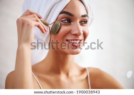 Close-up photo of a young smiley woman using a massaging roller on her face