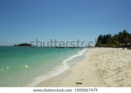 Photo of white sand turquoise water, some plants, people and small island  in the distance.