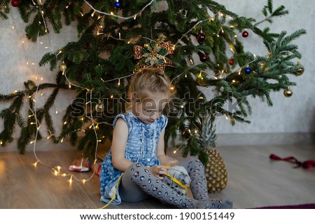 A small, pretty, blonde-haired, blue-eyed girl in a denim dress sits under a Christmas tree. There is a pineapple nearby