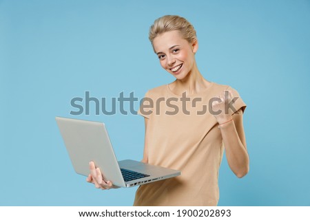 Young smiling blonde woman 20s with short haircut dental braces in casual beige t-shirt chat modern laptop pc computer doing winner gesture clenching fist isolated on blue background studio portrait