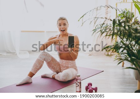 Amazed young strong sporty fitness woman wearing pink tracksuit doing yoga exercises sitting pointing index finger on mobile cell phone with blank empty screen stretching on mat floor at home indoor