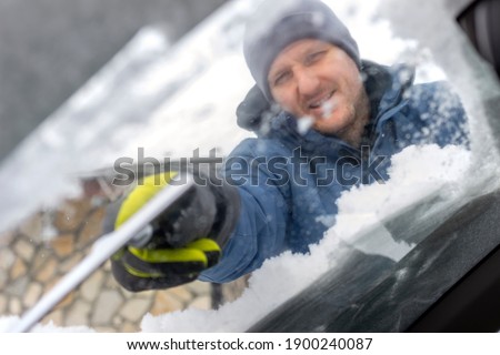 Photo of a young man cleaning snow covered car with a scraper. Close up of man remove snow from windshield. Shot of men cleaning car windshield from snow and ice after snow storm. Copy space.
