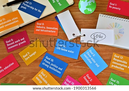 There is a table with a card with the SDG goals and a ball of earth, a small sketchbook with the SDG symbols and a word book with the letters of the SDGs. Royalty-Free Stock Photo #1900313566