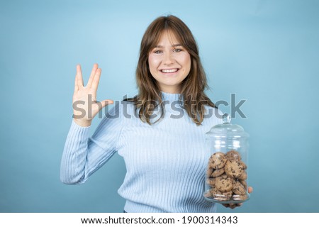 Young beautiful woman holding chocolate chips cookies jar over isolated blue background doing hand symbol