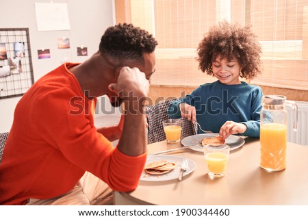 Healthy breakfast. Careful father asking something of his child politely. Cute curly boy with his father are sitting at the table and eating with pleasure emotions. Stock photo