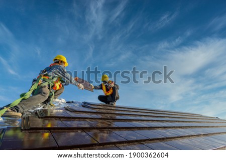 Two roofers Wearing safety clothing is working as a team to install the roof of the house that Ceramic tile roof at construction site Royalty-Free Stock Photo #1900362604