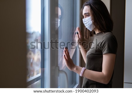 Sad young woman in a medical mask looks out of the window. Quarantine during the coronavirus pandemic. Royalty-Free Stock Photo #1900402093