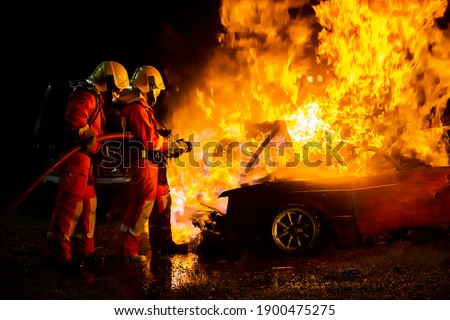 Firefighter in fire fighting suit spraying water, Firemen fighting raging fire with huge flames of burning car, Fire prevention and extinguishing concept
