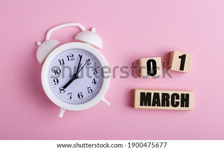 On a pink background, a white alarm clock and wooden cubes with the date of MARCH 01