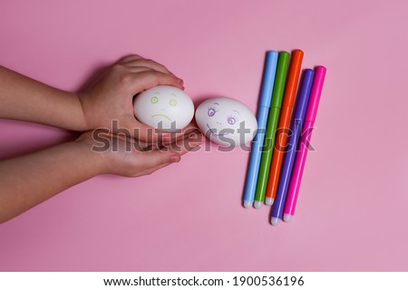 Children's hand holds two eggs sad and cheerful. Easter, draws emoticons, faces, emotions. Food markers, creativity. Pink background.