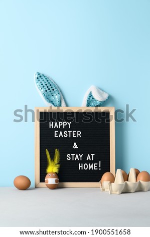 Easter holiday concept 2021. Easter bunny made from egg wearing protective face mask, and black felt letter board with slogan - Happy Easter and Stay at Home on pastel cyan background