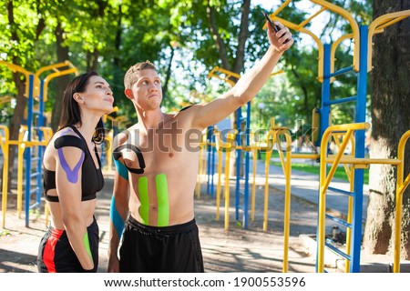 Professional caucasian athletes with kinesiology elastic taping on bodies, handsome man and brunette woman, posing at sports ground, having rest and taking selfie using smartphone.
