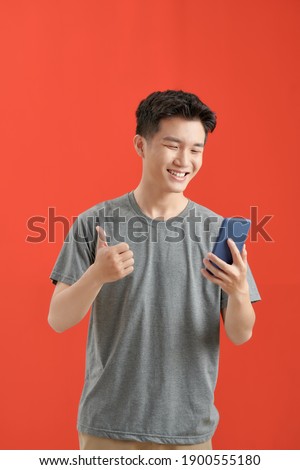 Portrait of a young cheerful excited bearded man wearing t-shirt standing isolated over red background, using mobile phone, showing thumbs up