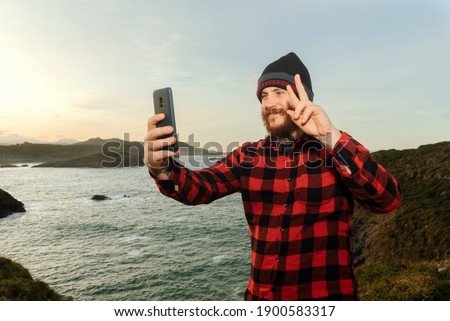 Smiling bearded man taking selfies making the victory sign with his smartphone during a trip along the coast of Asturias, Spain. Person using his mobile phone.