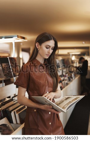 Attractive stylish young caucasian woman in brown dress, reading magazine, turn pages in book with focused, interested expression, picking what buy in vintage store, contemplate pictures.