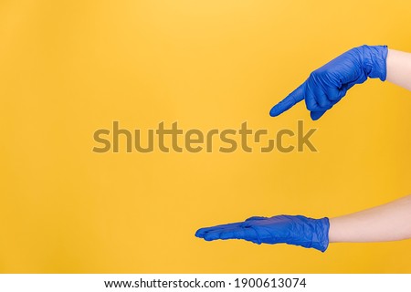 Unrecognizable young female opening and pointing on palm on copy space aside, wears medical protective blue gloves, isolated on yellow studio background for advertisement. Body language concept